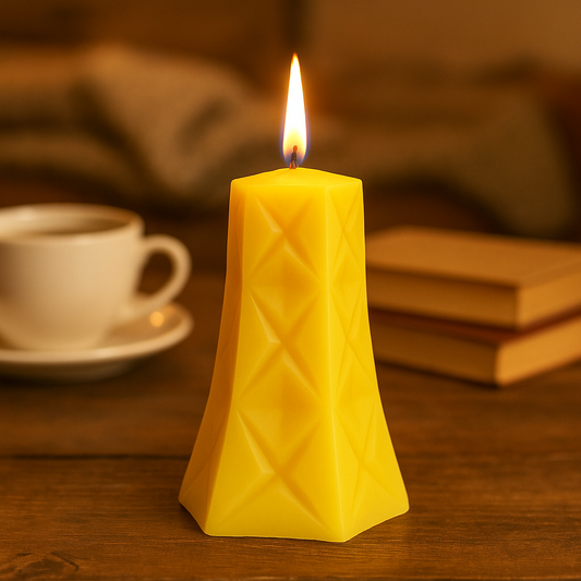 Yellow diamond patterened pillar candle with a burning wick on a wooden surface with a cup and books in the background.